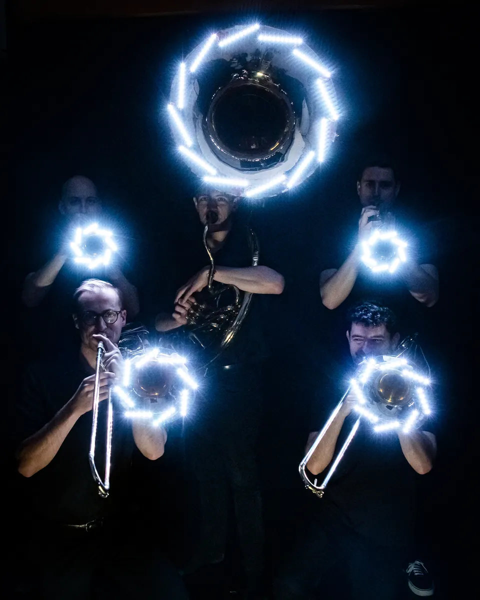 Brass players hold bright white LED-lit instruments in a dark portrait.