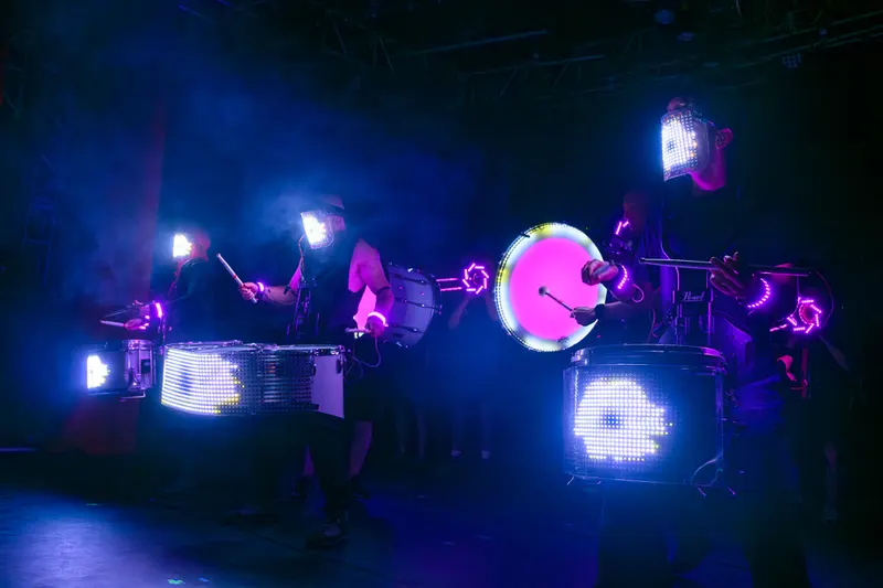 Band members in LED visors perform behind illuminated drums.